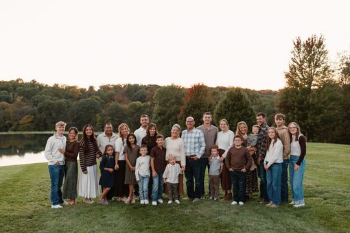 A large multi-generational family poses for a portrait in a sunlit, grassy field during the fall.