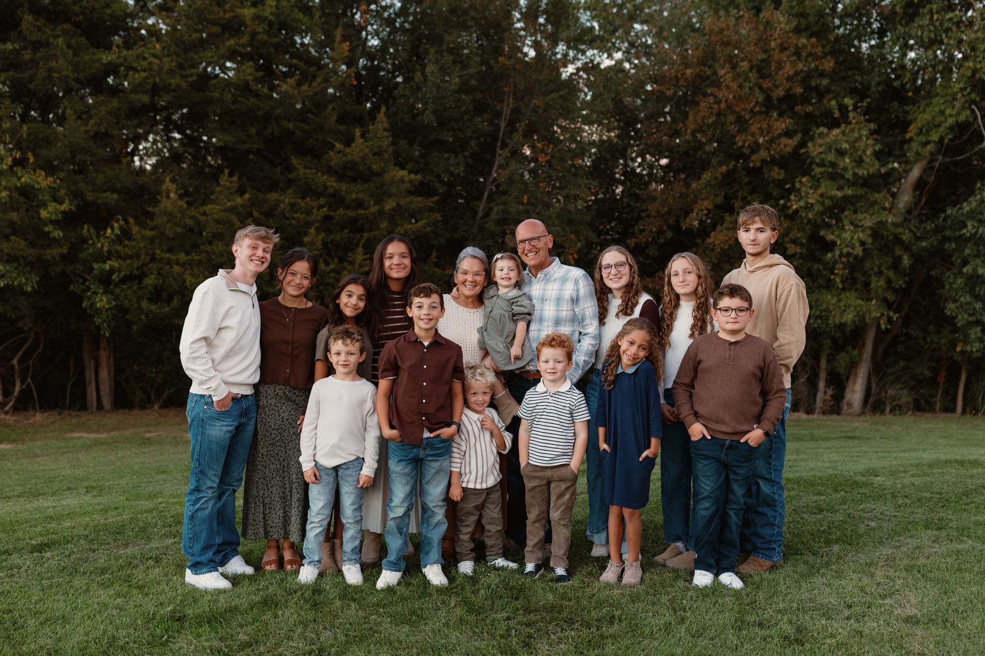 A large family posing outdoors in a grassy field during golden hour, with trees in the background.