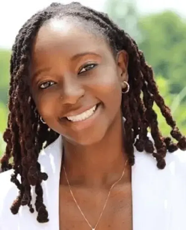 Smiling person with braided hair and hoop earrings outdoors, wearing a white top.