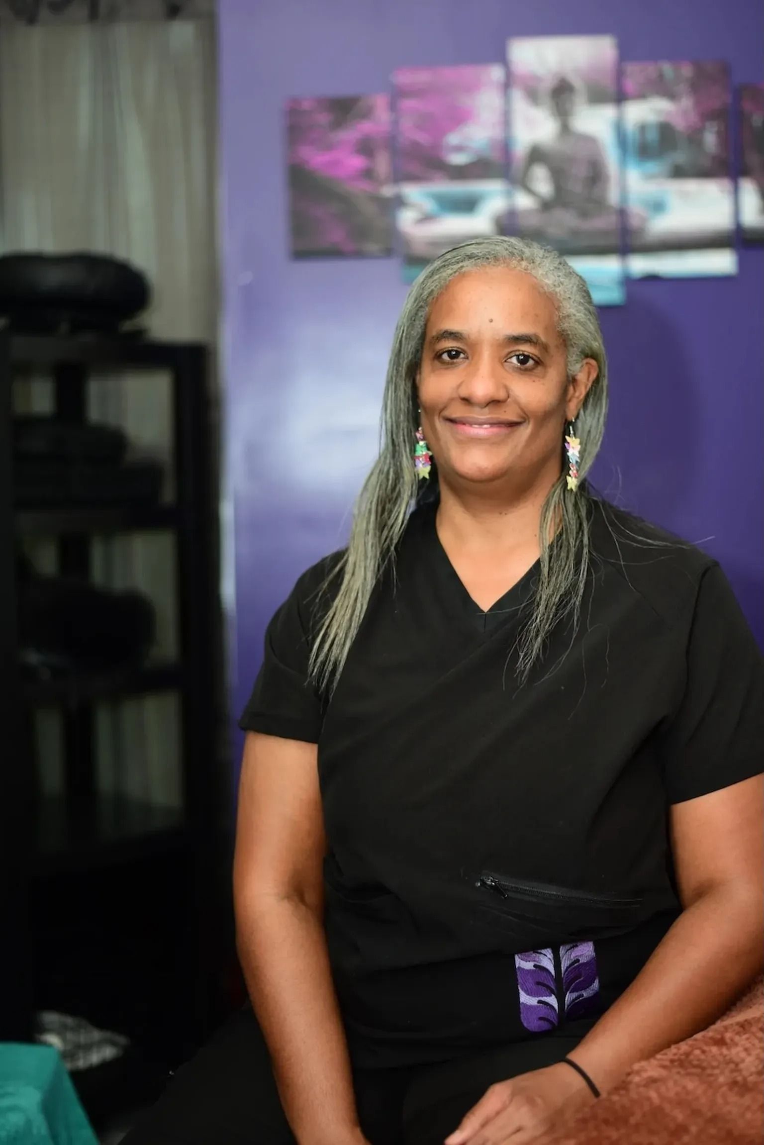 Portrait of a smiling person with gray braided hair, seated against a purple studio backdrop