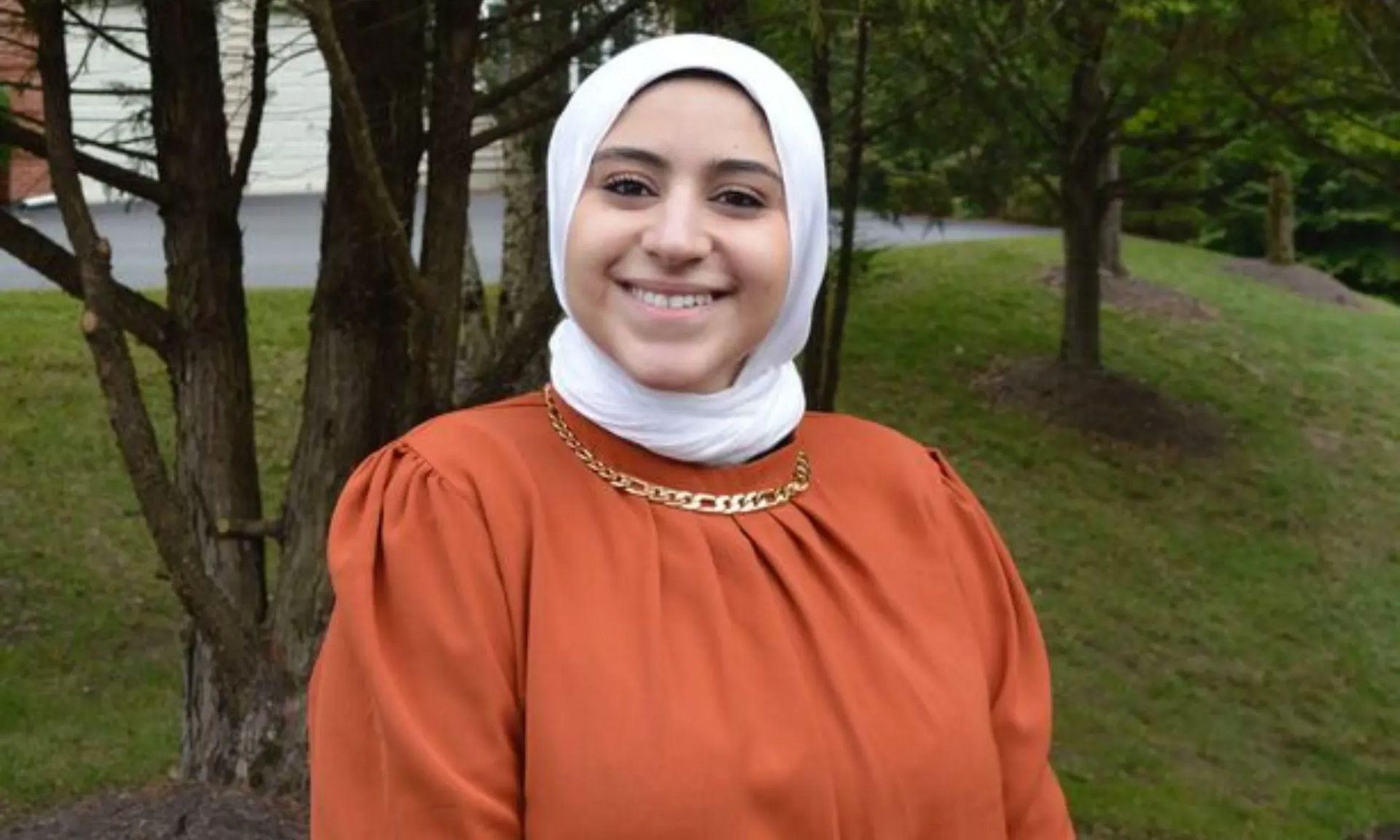 Smiling person in a white headscarf and orange top standing outdoors near trees and grass