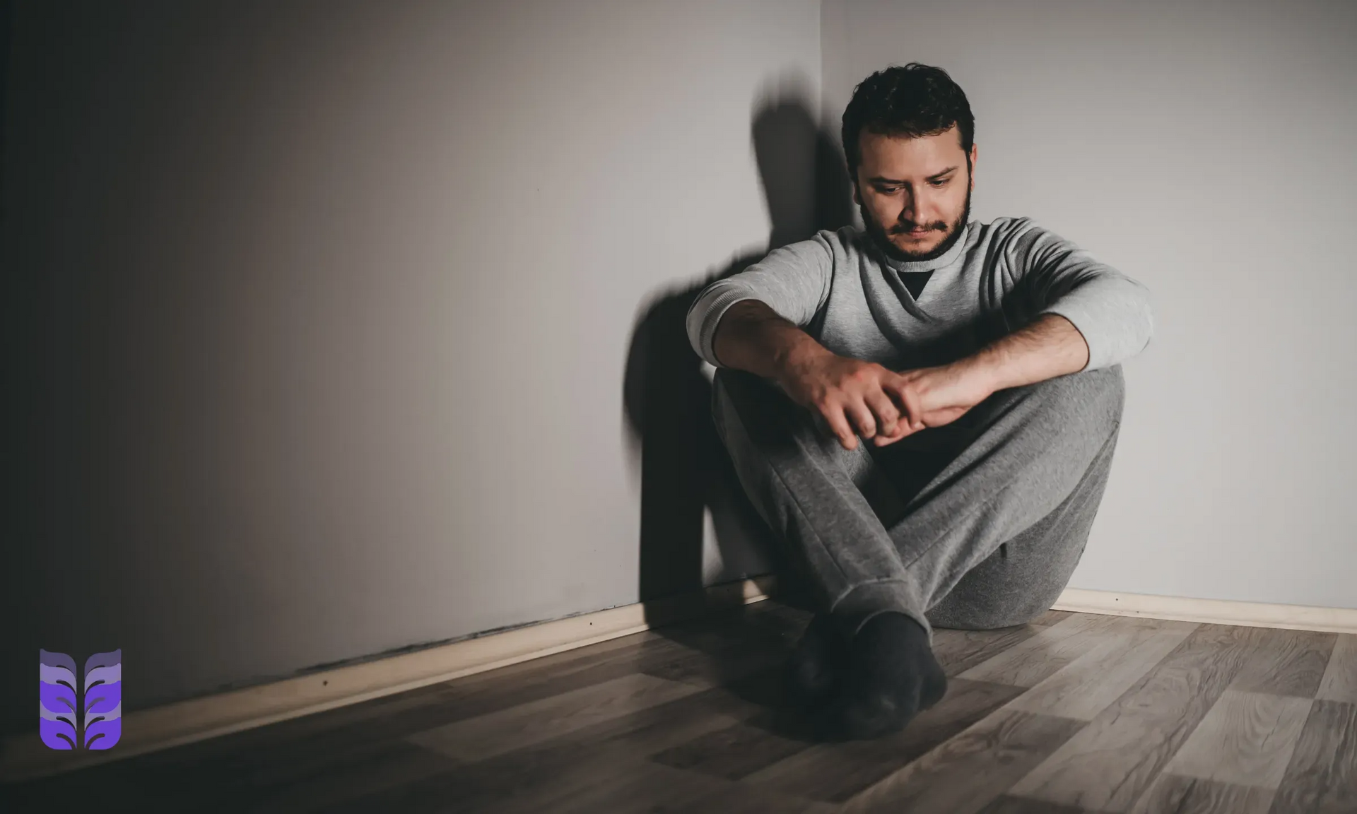 Man sitting on the floor against a wall, looking down, in a dim room with a wooden floor