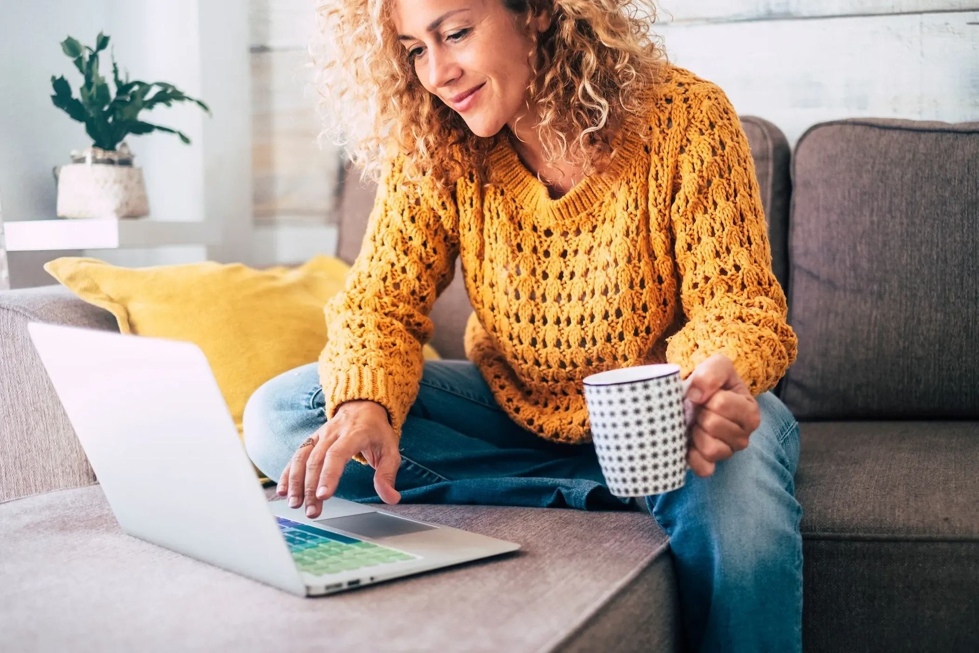 Woman on a couch using a laptop and holding a mug in a bright living room