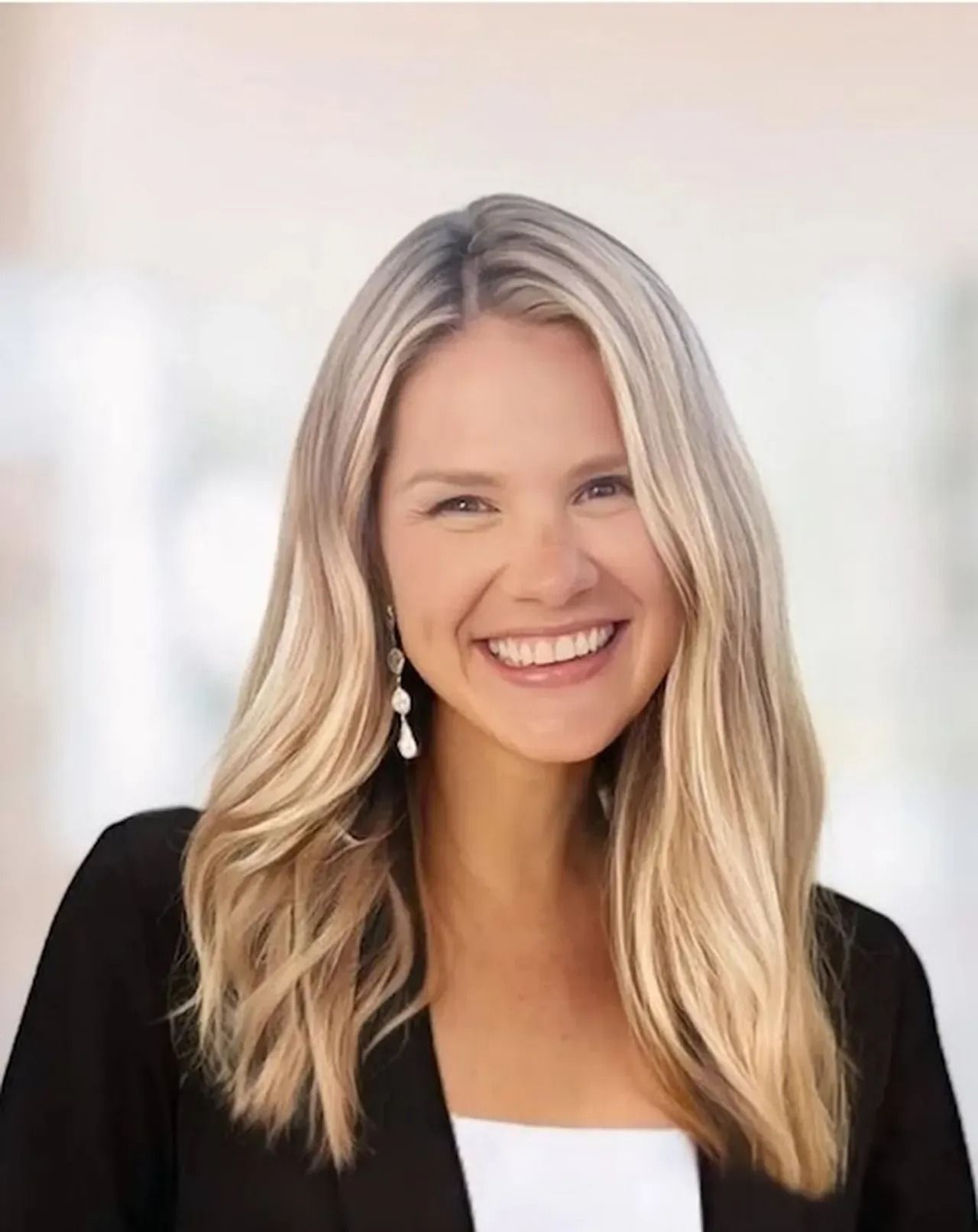 Smiling woman with long blonde hair wearing a black blazer and white top against a soft white background