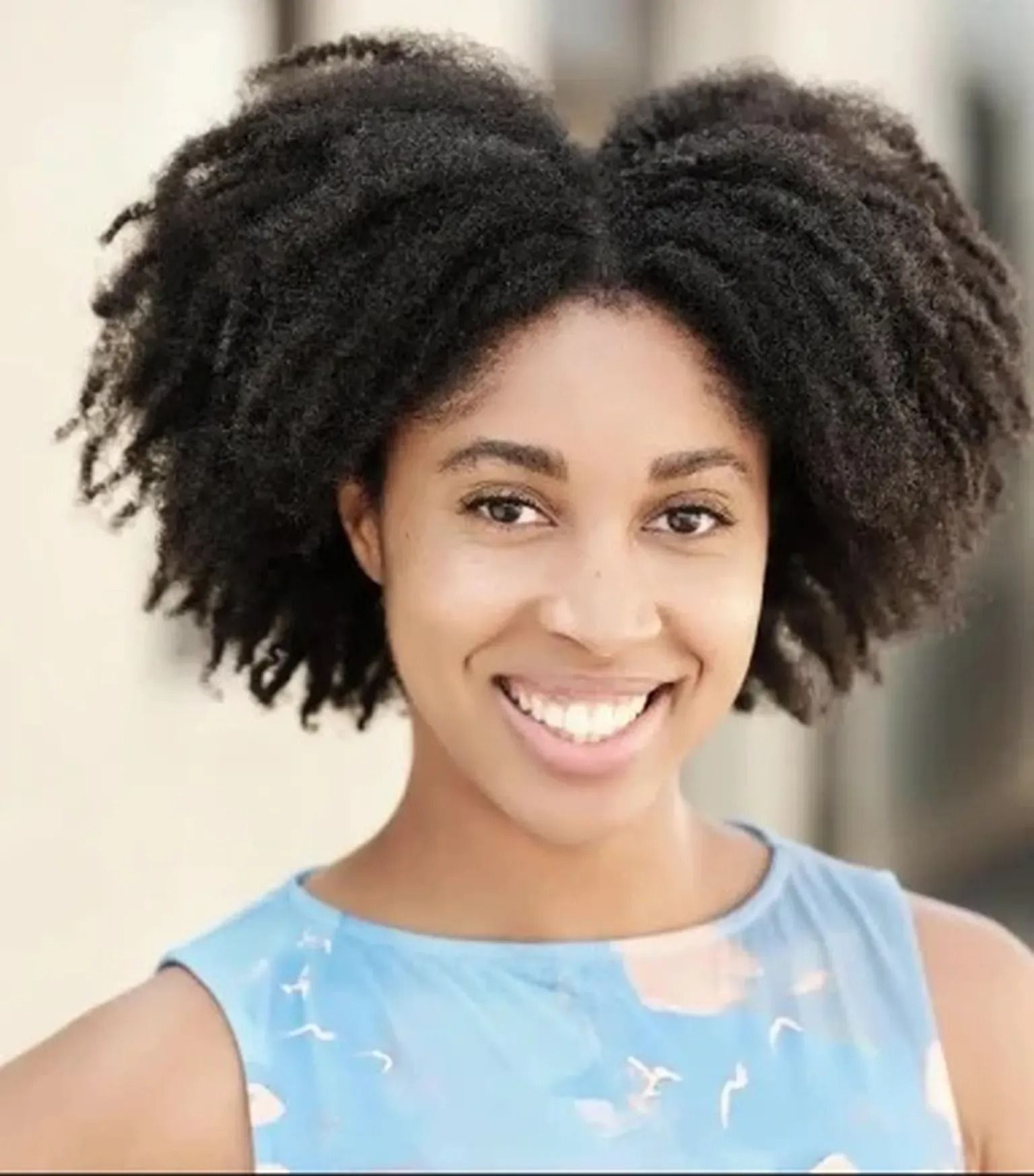 Smiling person with natural curly hair wearing a light blue sleeveless top, against a soft blurred background.