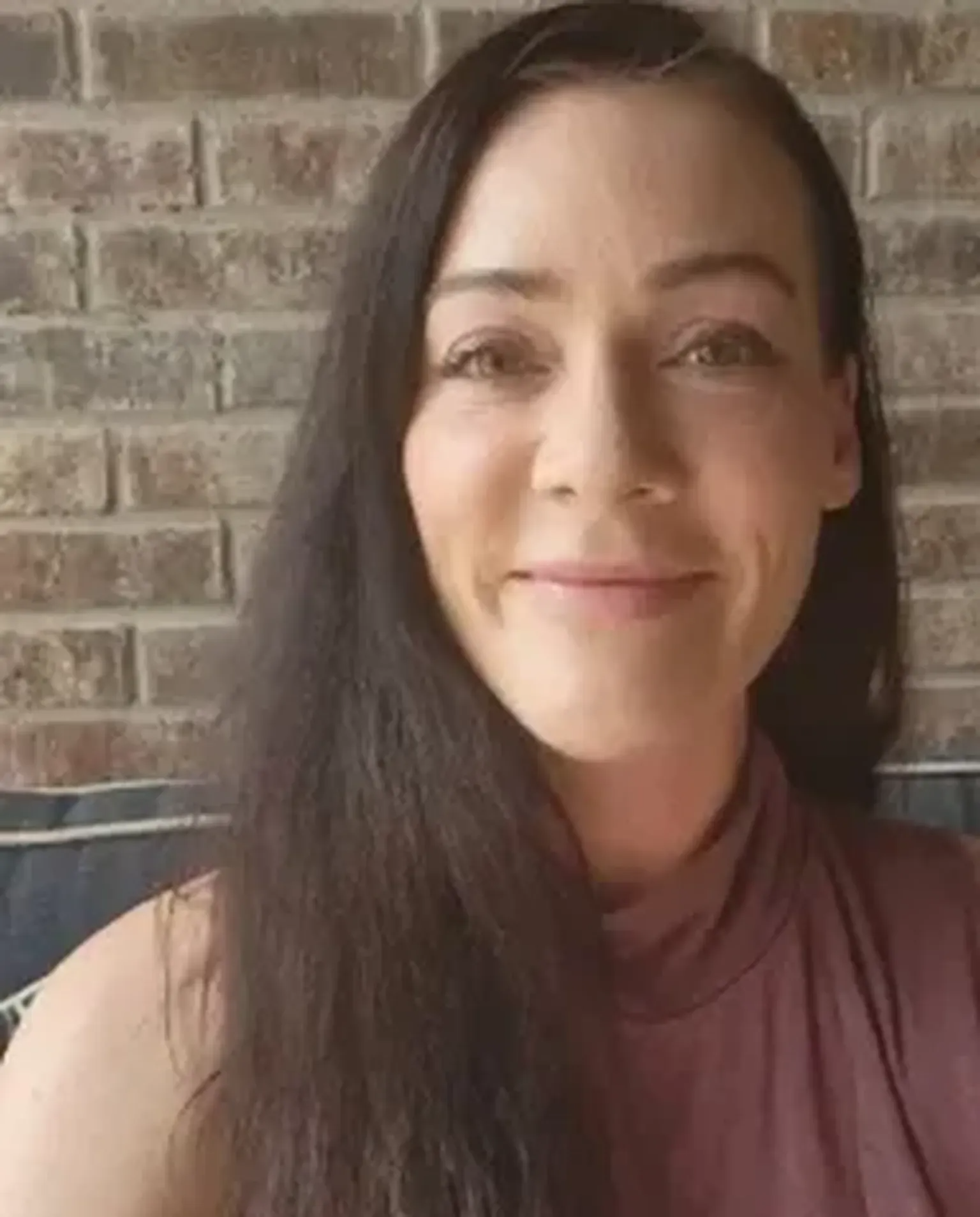 Smiling person in a mauve top seated against a brick wall background