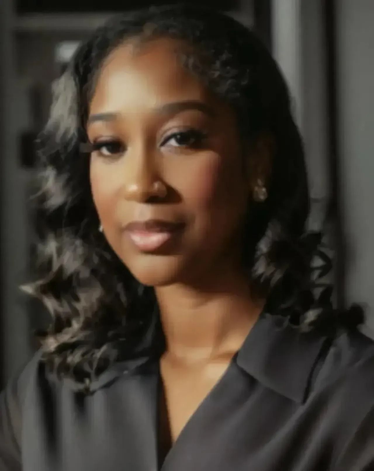 Portrait of a person in a dark blouse, softly lit against a blurred gray background, with a calm expression