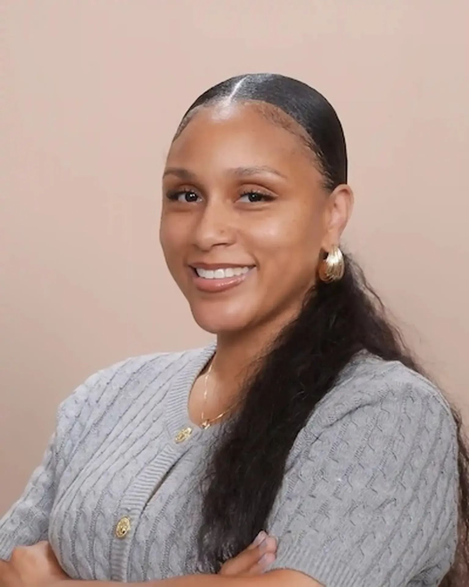 Smiling woman with long dark hair, wearing a gray sweater and gold jewelry against a beige background