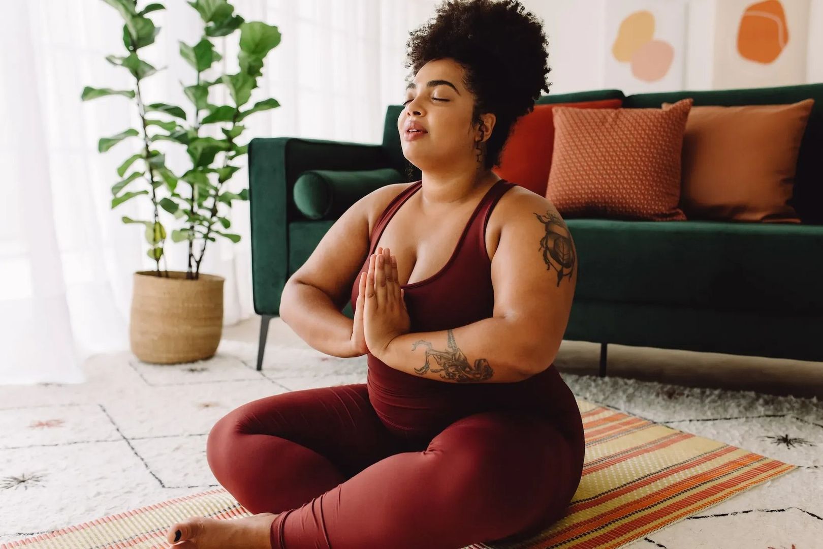Person meditating on a yoga mat in a bright living room, wearing burgundy workout clothes.