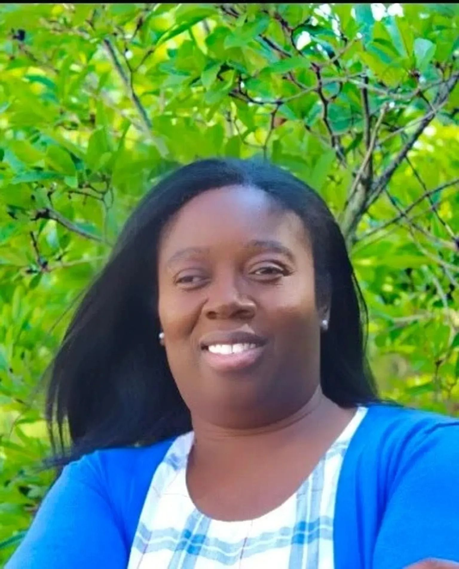 Smiling woman in a blue top standing outdoors with green leafy branches behind her