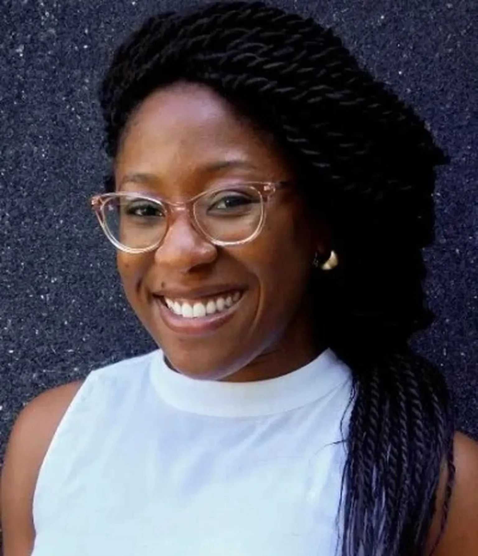 Smiling person with glasses and braided hair wearing a white top against a dark background