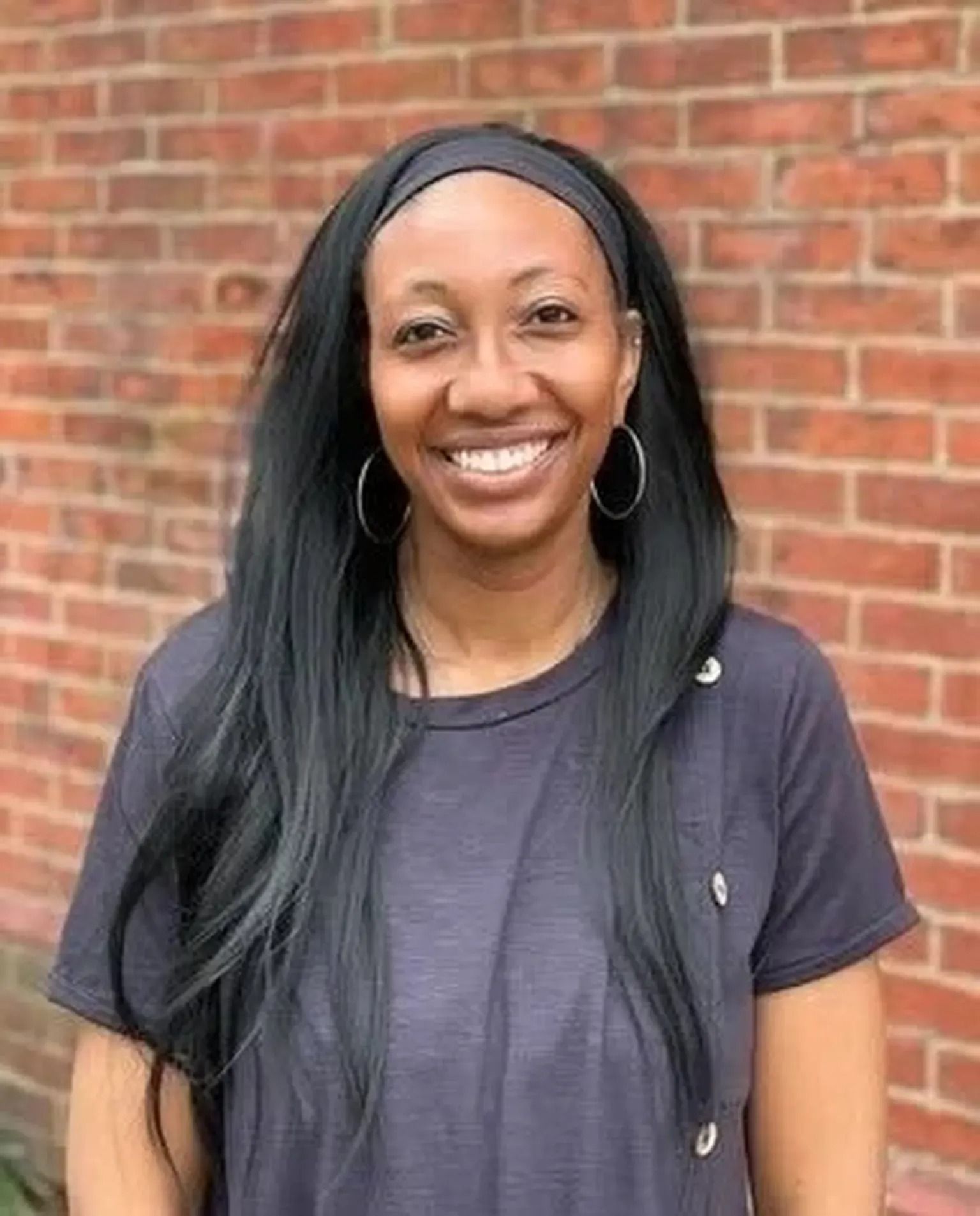 Smiling woman with long black hair, wearing a dark T-shirt, standing in front of a brick wall.