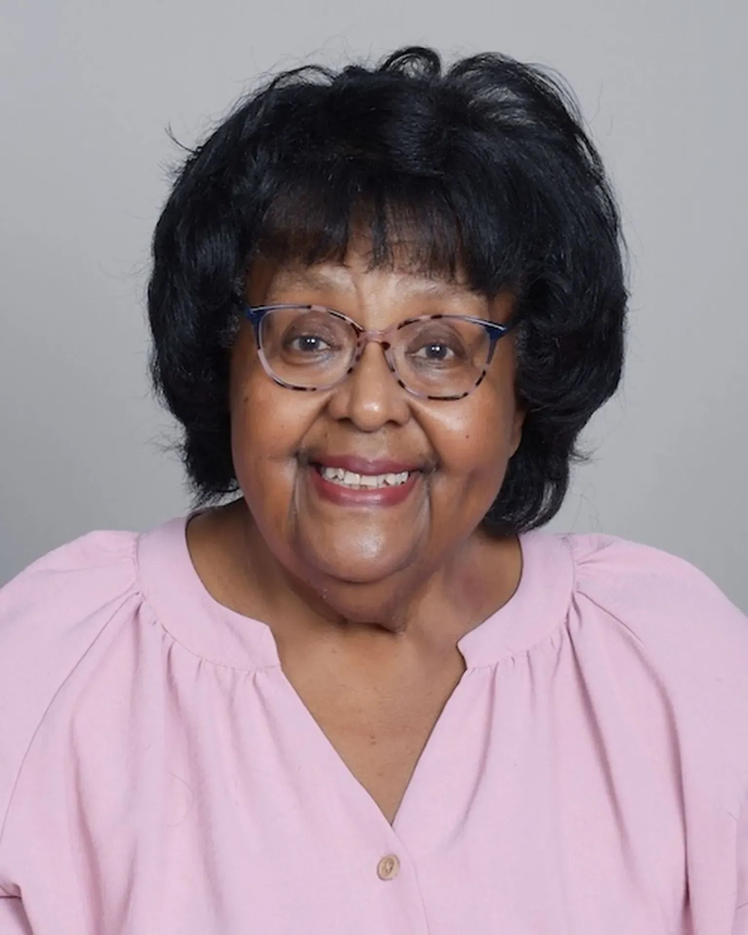 Smiling woman with glasses and black hair, wearing a light pink blouse on a gray background