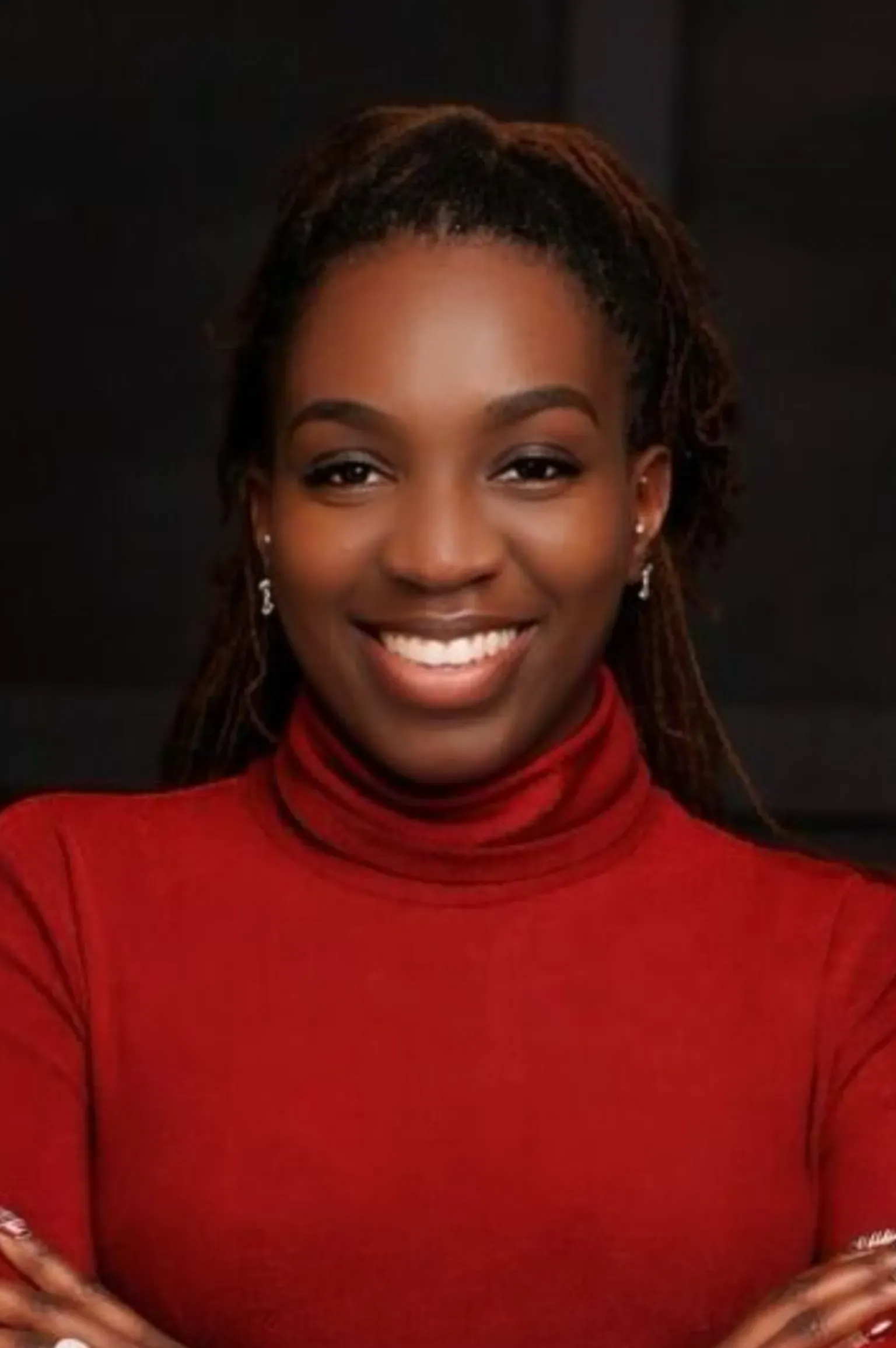 Smiling person in a red turtleneck against a dark background, arms crossed.