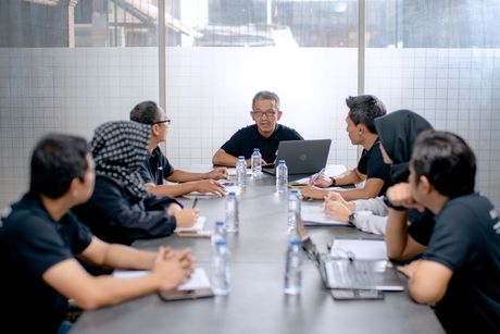 Team meeting around a conference table with laptops and water bottles, led by a speaker at the center.
