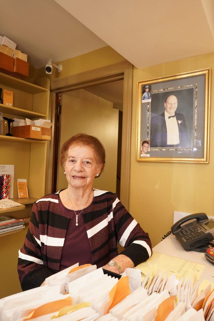 A person in a striped sweater sits behind a desk in an office with shelves and a framed portrait on the wall.