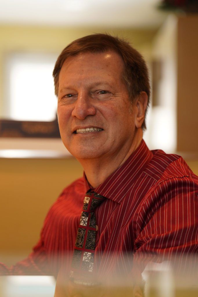 A smiling man wearing a red pinstriped button-down shirt and a patterned tie, indoors against a blurred background.