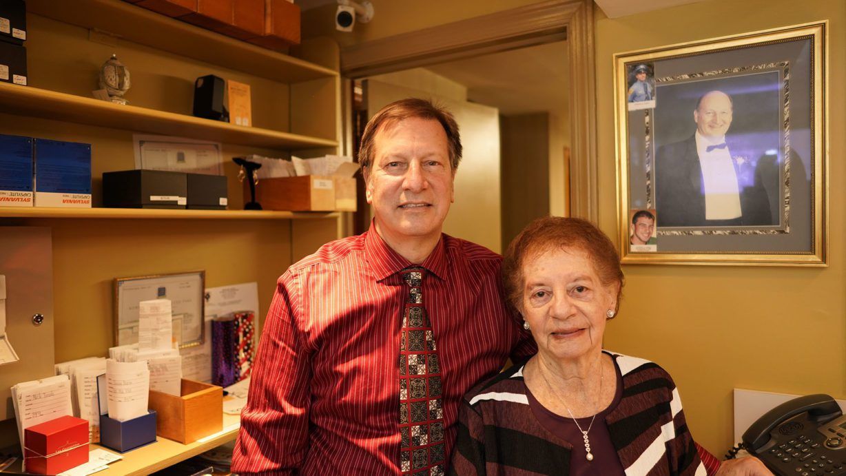 Two people stand in an office, one wearing a patterned tie, beside a framed portrait of a man on a tan wall.