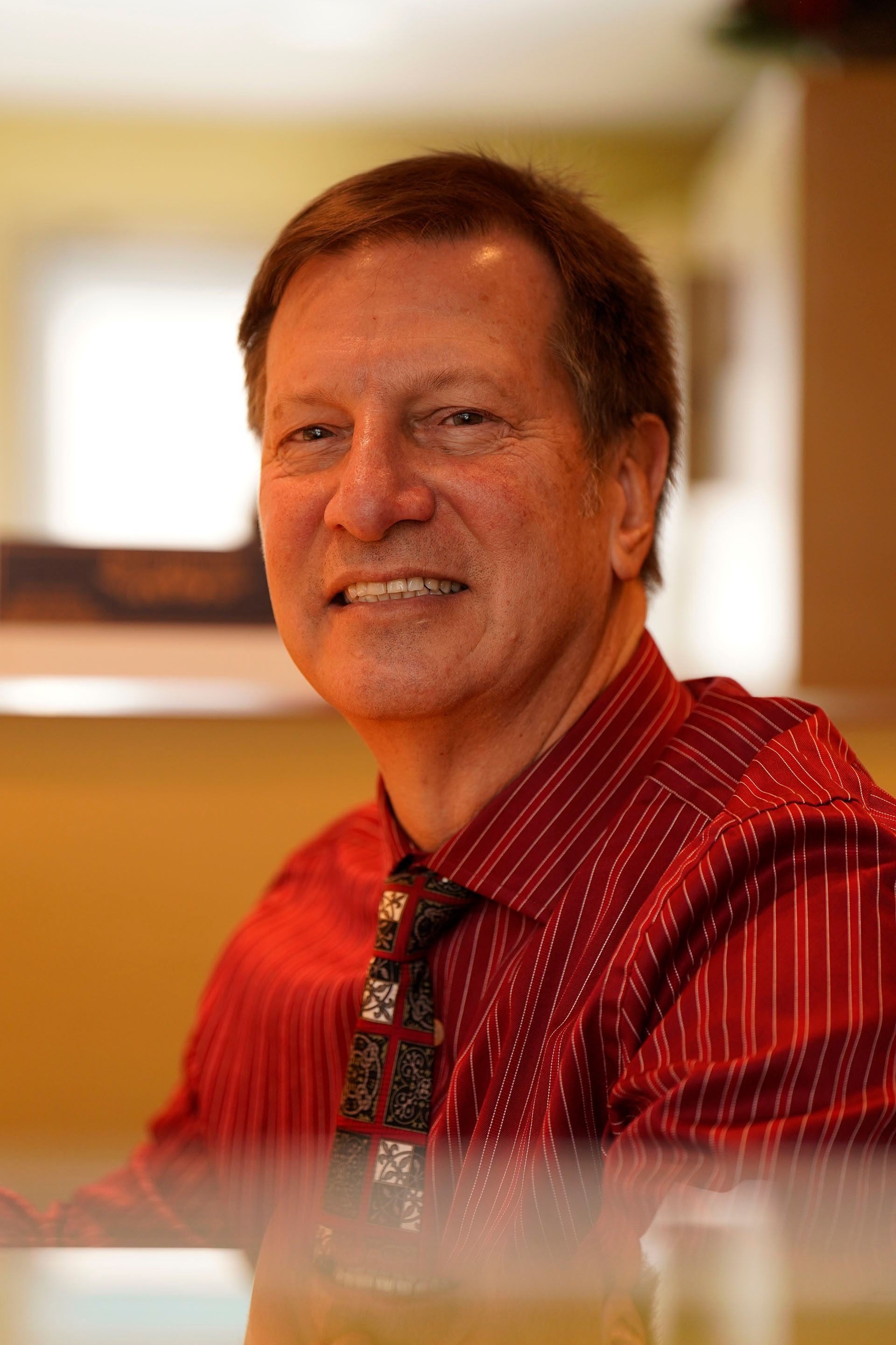 A person wearing a red pinstriped button-down shirt and a patterned necktie, smiling in a softly lit indoor setting.
