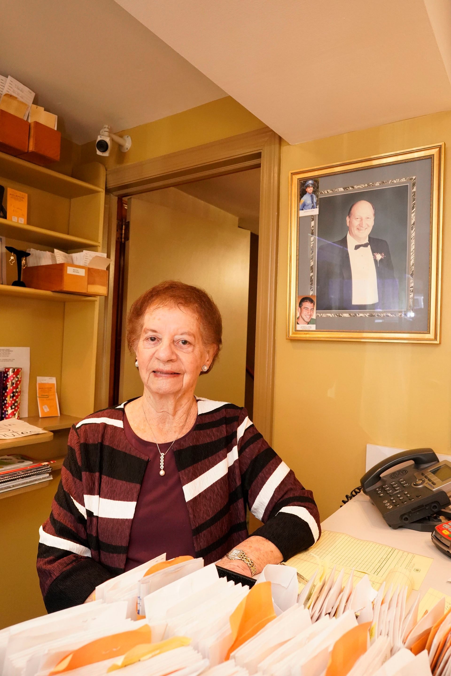 A person in a striped cardigan sits at a desk with organized files, with a framed portrait on the yellow wall behind.