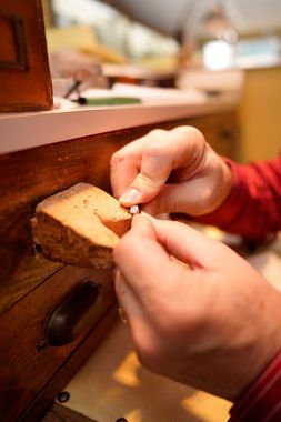 A jeweler’s hands work on a small, glittering ring at a wooden workbench with a stone bench pin.