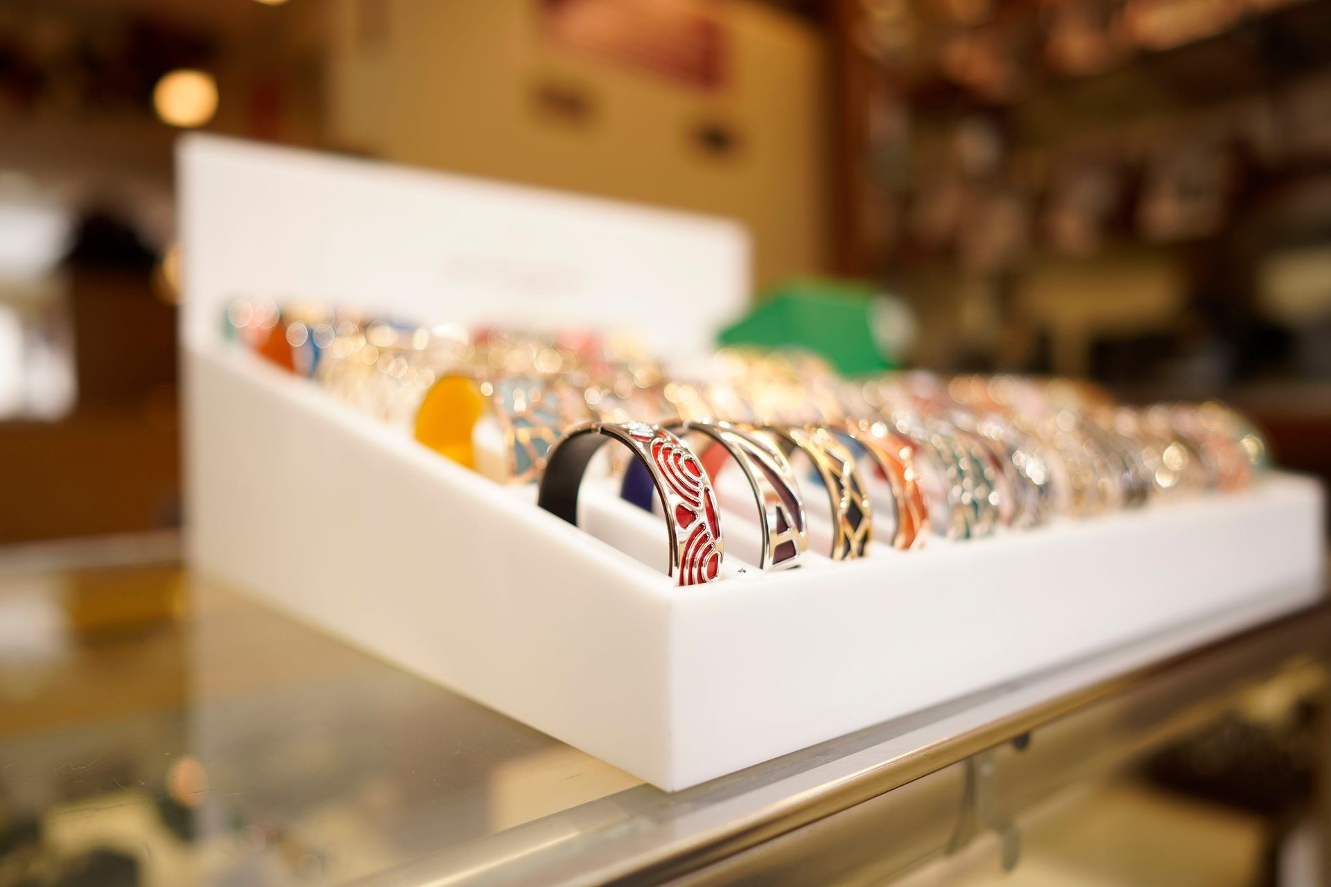 A slanted white display case on a glass counter features a variety of colorful patterned bangle bracelets.