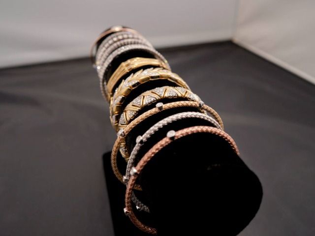 A row of various metallic bracelets and bangles in gold, rose gold, and silver tones arranged on a dark display stand.