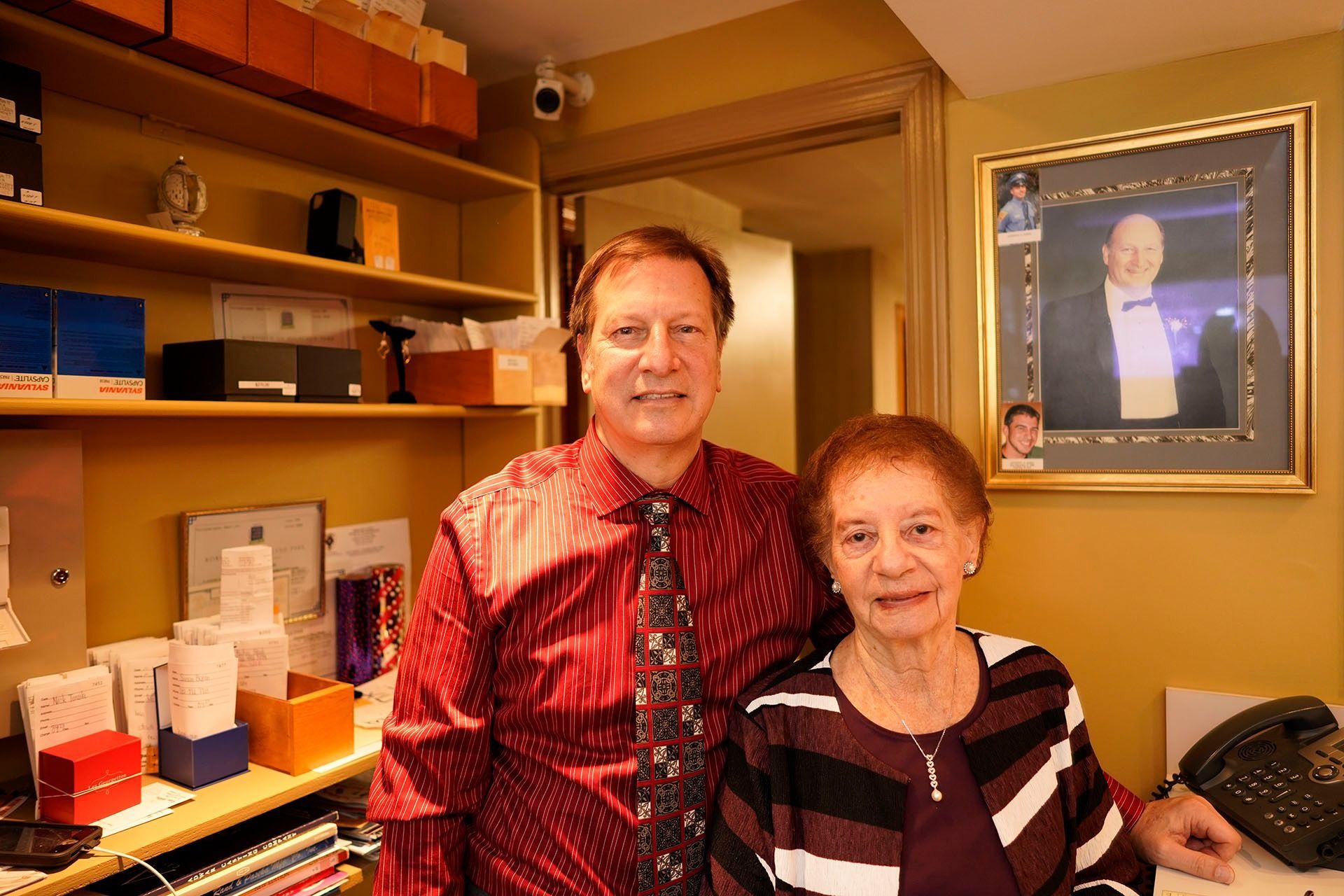 A man in a red shirt and patterned tie stands beside a woman in a striped top in a room with shelves and a framed photo.