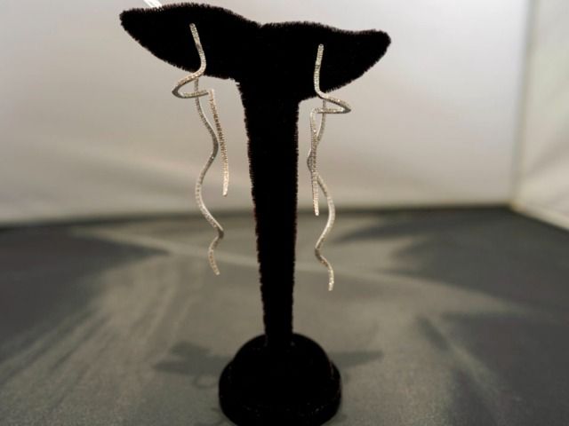 Two long, thin, wavy silver earrings hang from a black velvet T-bar display stand against a blurred background.