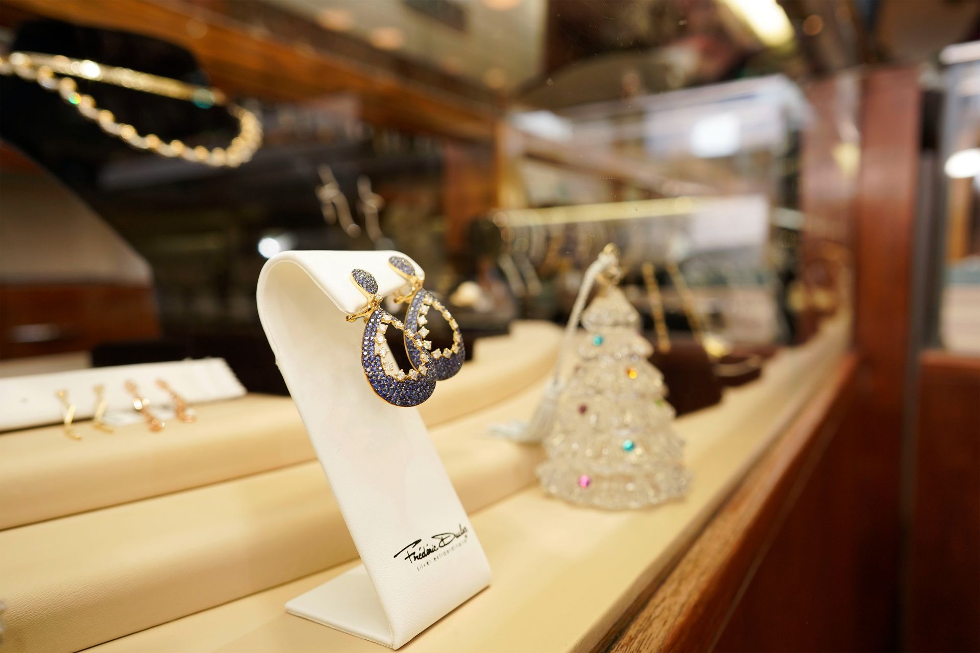 A pair of gold earrings with blue stones displayed on a stand in a jewelry store case next to a small glass tree.