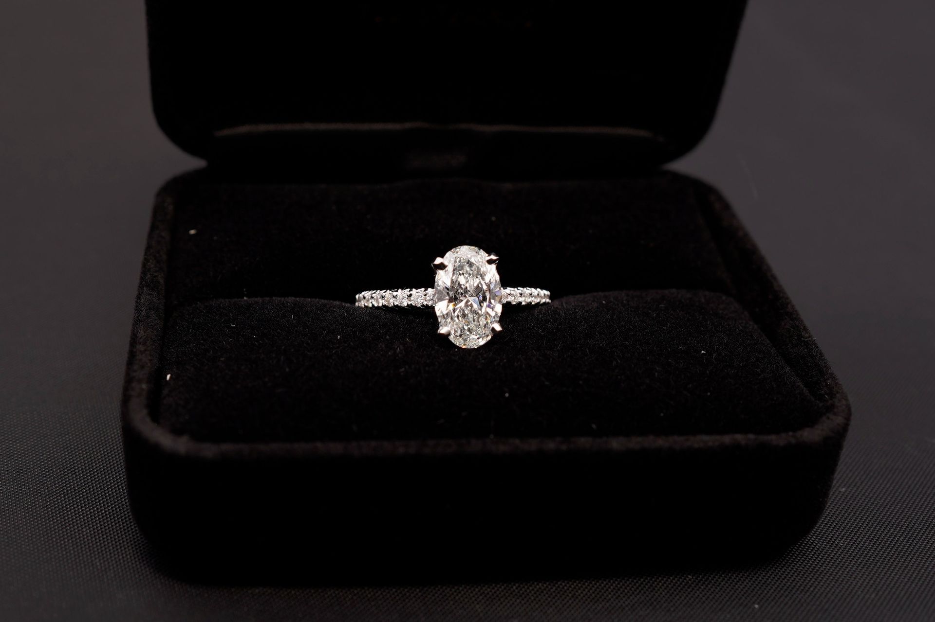 An oval-cut diamond engagement ring with a pavé band set inside an open, black velvet jewelry box.