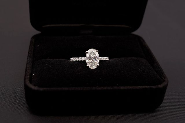 A diamond engagement ring with an oval-cut stone and a pavé band, centered inside a black velvet ring box.