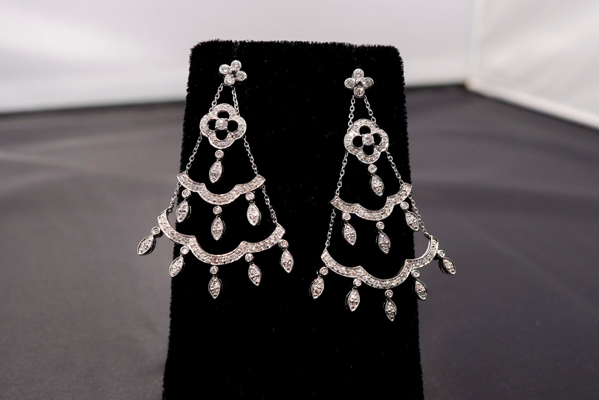 A pair of chandelier-style silver earrings featuring floral accents and cascading, teardrop-shaped diamonds on a black stand.