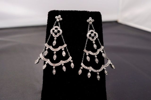 A pair of silver-toned, multi-tiered chandelier earrings with dangling diamond-like stones, displayed on a black stand.