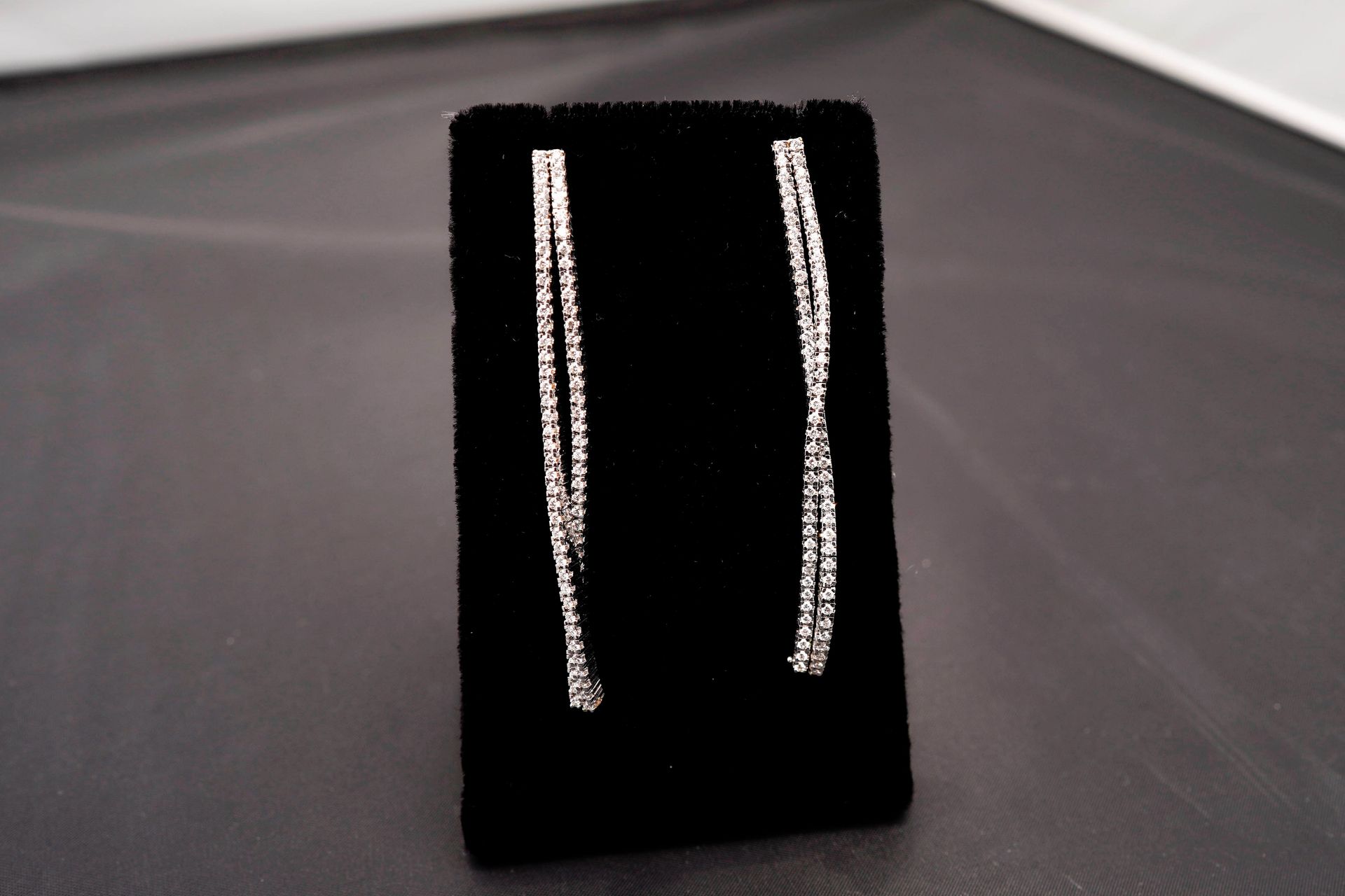 Two dangling, crystal-encrusted line earrings displayed on a black velvet stand against a gray background.