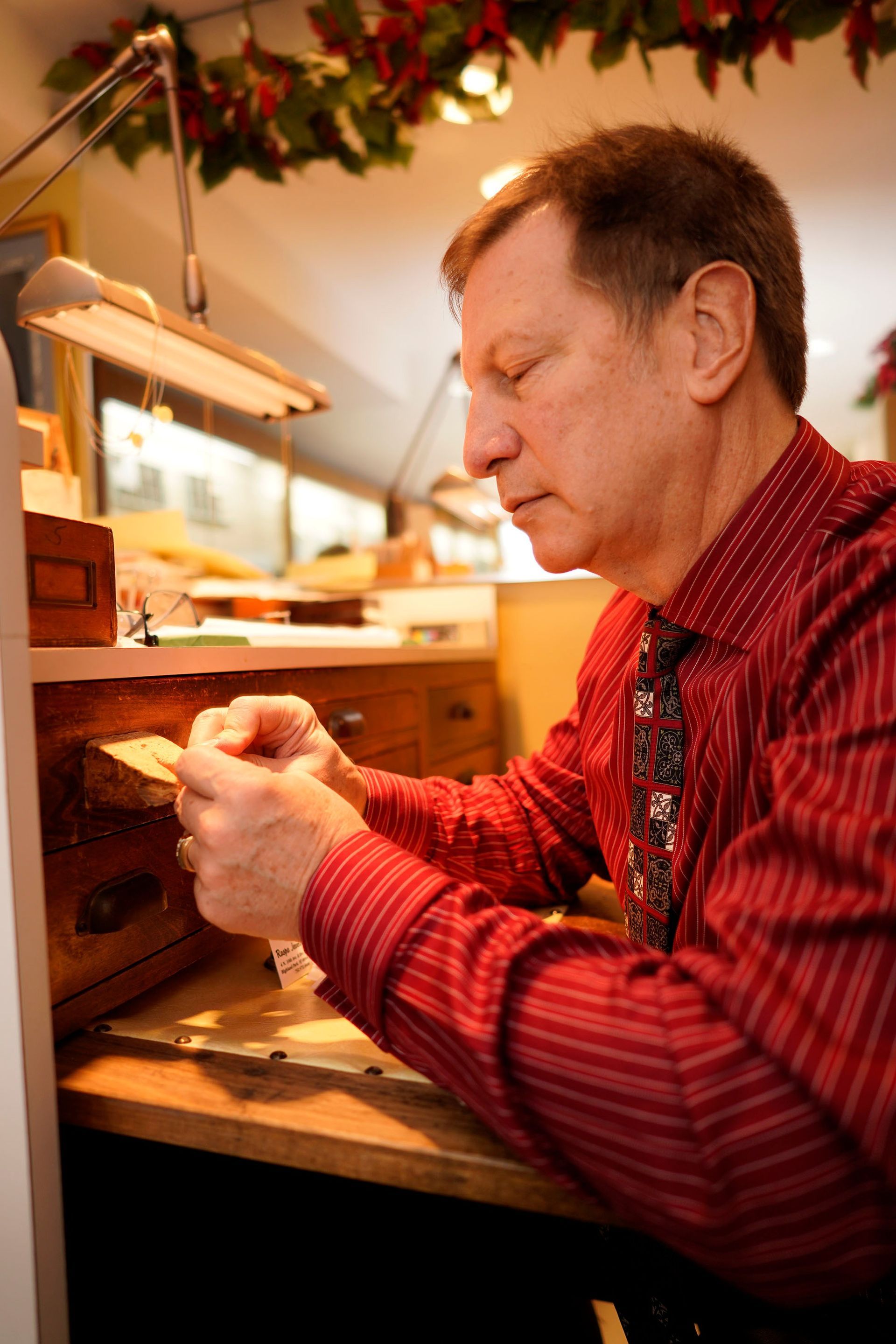 A person in a red striped shirt and patterned tie works at a desk, carefully holding a small object under task lighting.