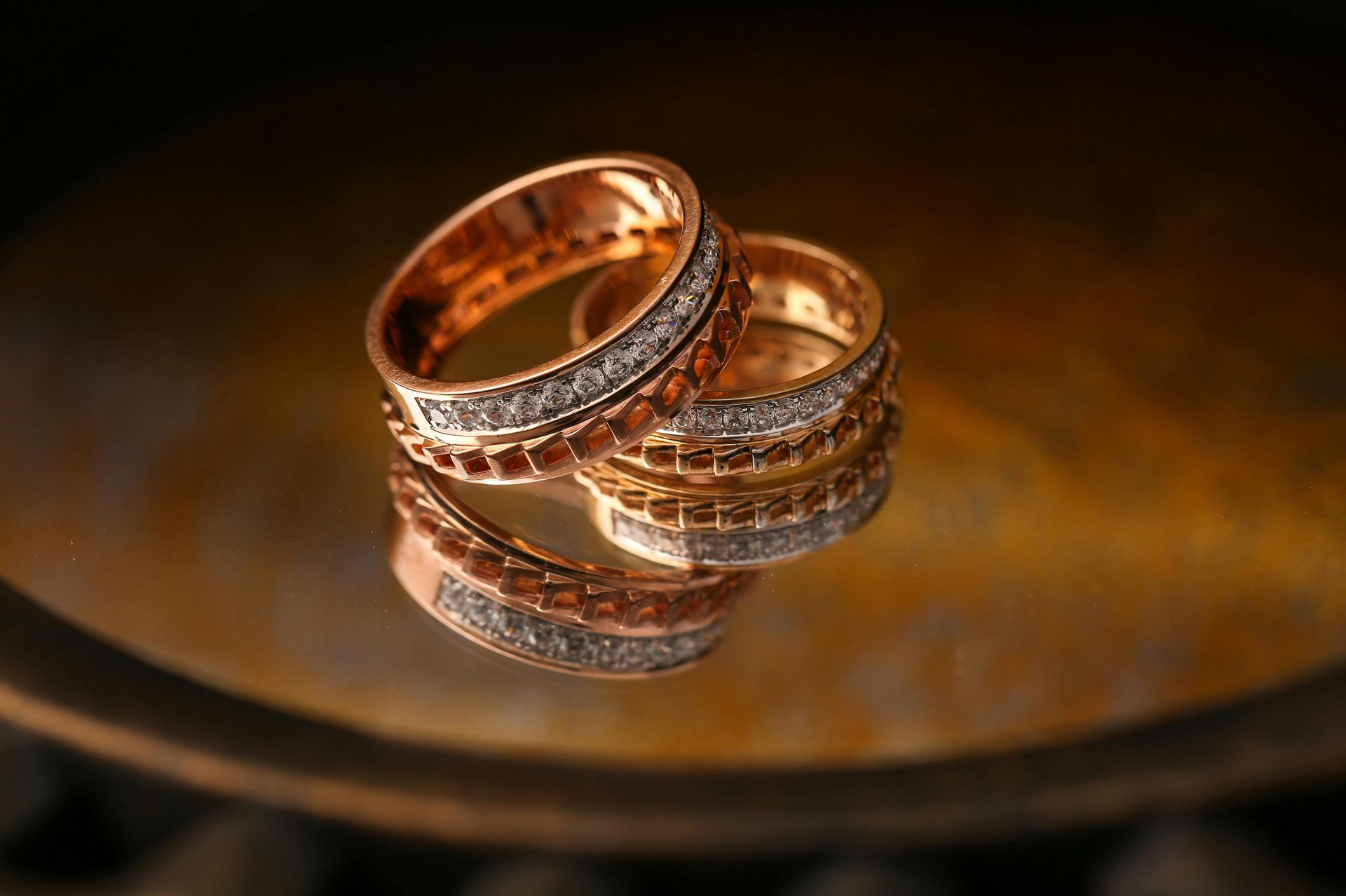 Two rose gold wedding bands with inlaid diamonds resting on a reflective, circular surface with a warm, golden glow.