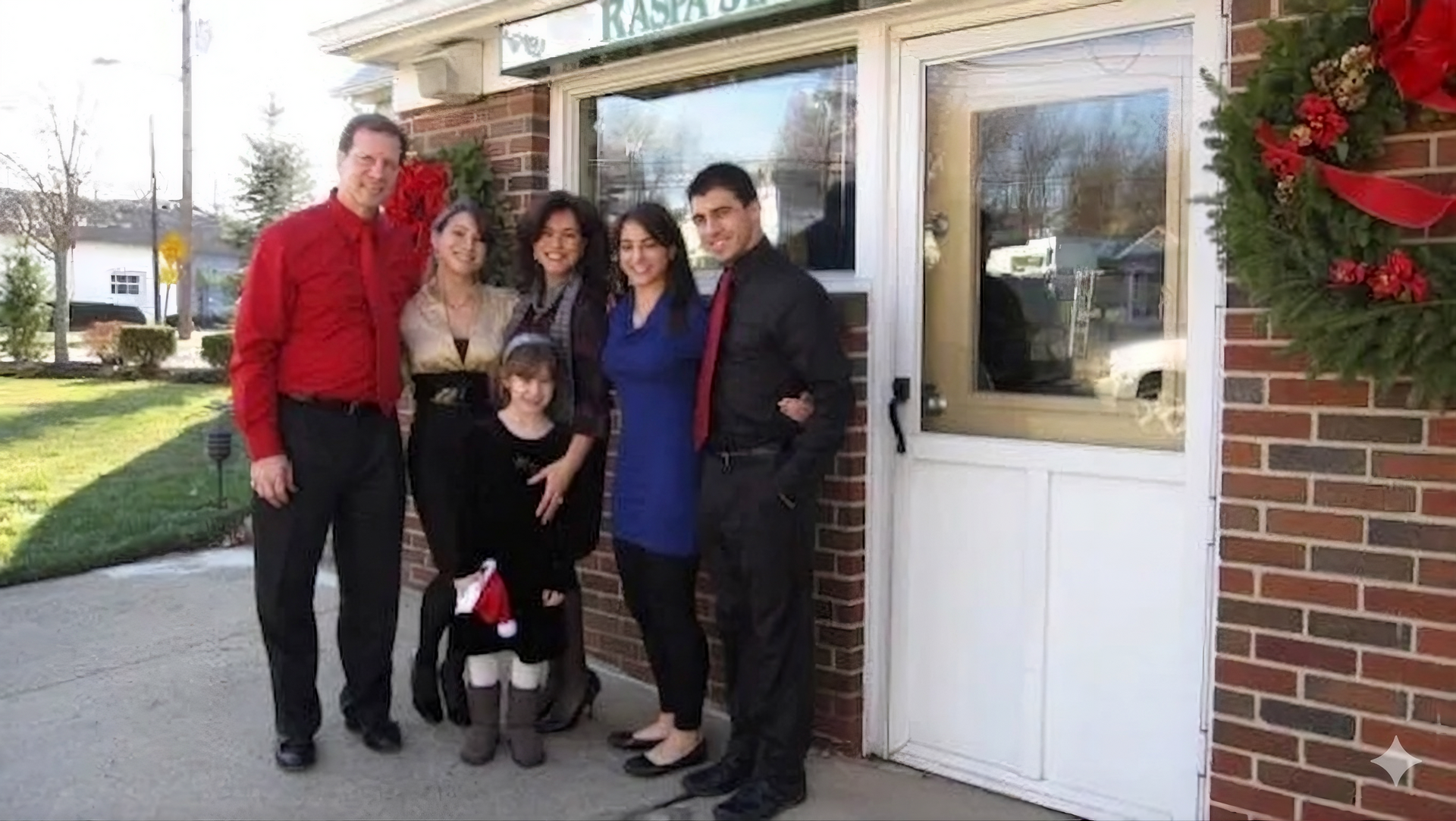 A group of six people stand smiling outside a brick building decorated with a holiday wreath.