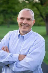 Smiling man in a light blue shirt with arms crossed, standing outdoors with a blurred green background