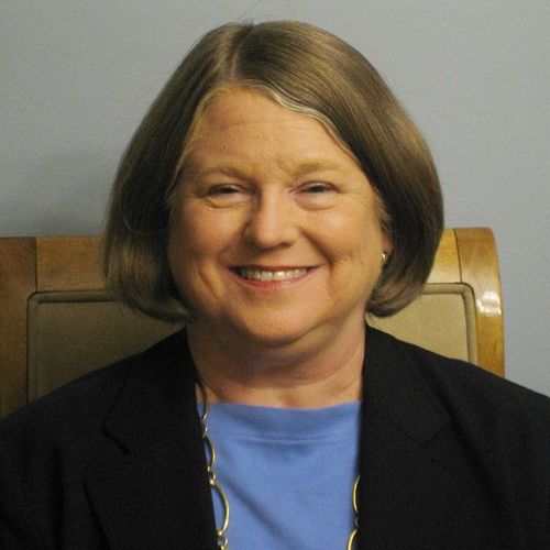 Smiling woman seated against a gray wall, wearing a black blazer over a blue top.