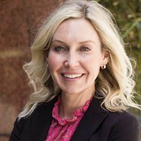 Smiling blonde woman in a black blazer and pink blouse against a blurred brick background