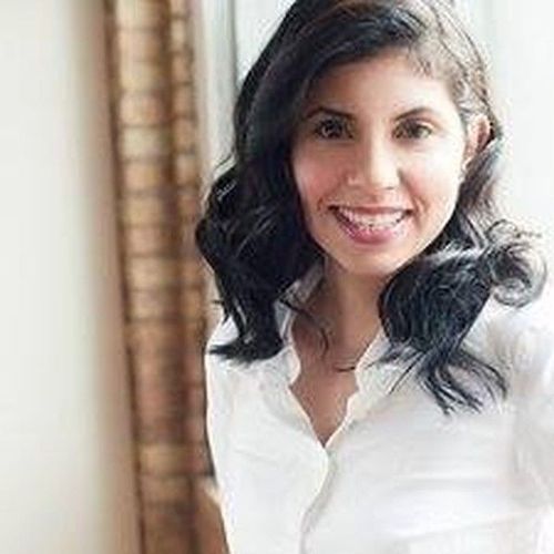 Smiling woman in a white blouse with dark curly hair, standing indoors by a window.