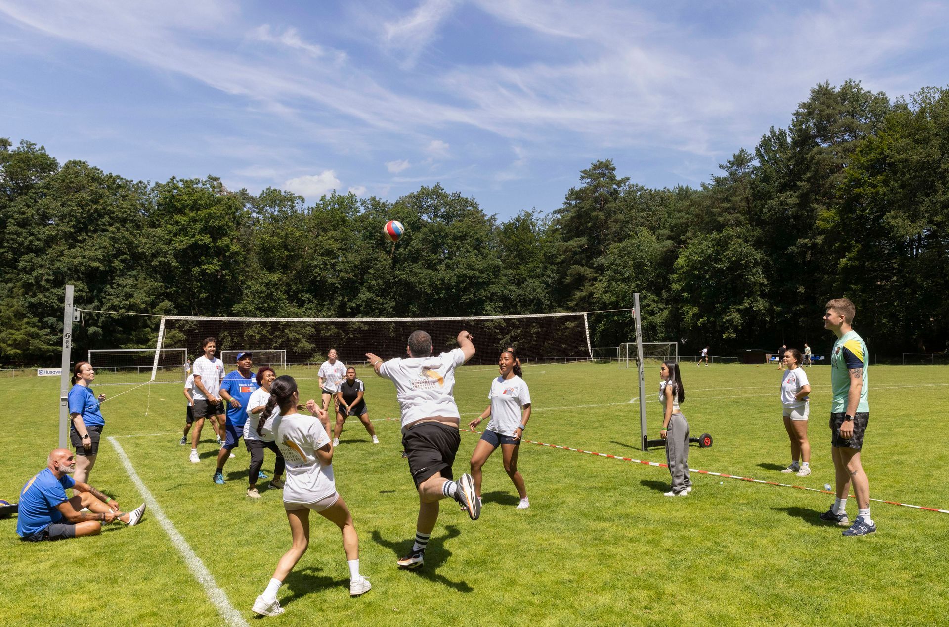 Een groep mensen speelt volleybal op een grasveld, met bomen op de achtergrond onder een blauwe, zonnige hemel.