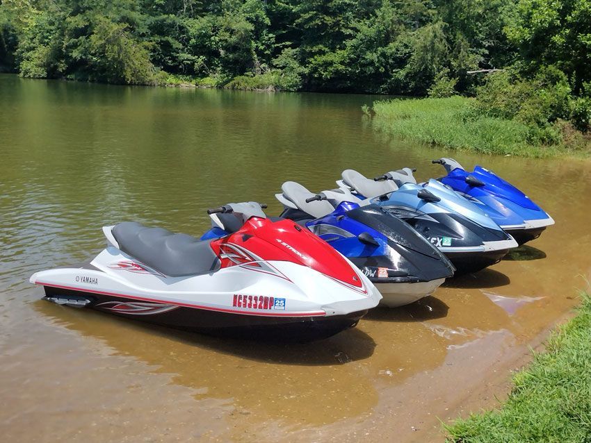 Four colorful jet skis lined up on a muddy lakeshore beside calm water and green trees