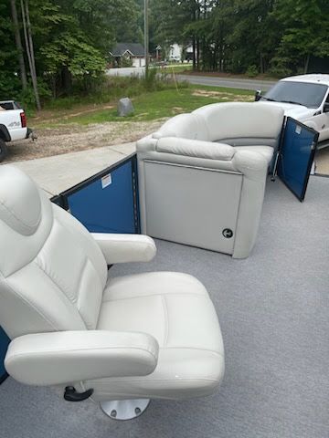 Two white leather recliners on a trailer outdoors, with blue side panels and a wooded driveway in the background