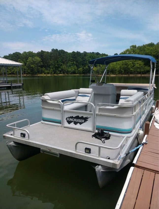 Pontoon boat docked beside a wooden pier on a calm lake, with blue canopy and gray seating.