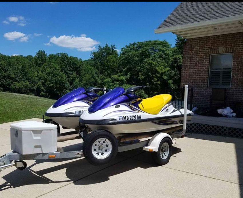 Two jet skis on a trailer in a driveway beside a house, one blue and white, one yellow and white.