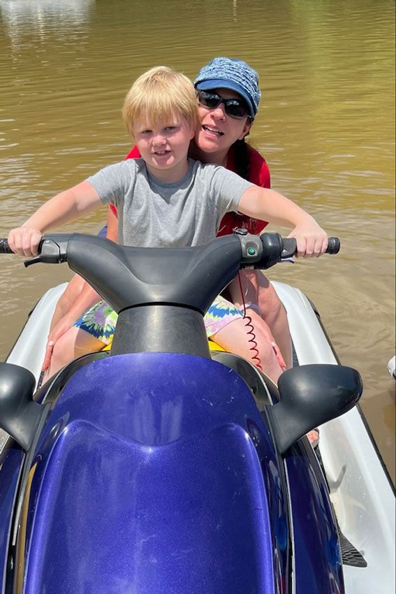 Two smiling children on a purple jet ski in muddy water, one wearing sunglasses and a blue hat