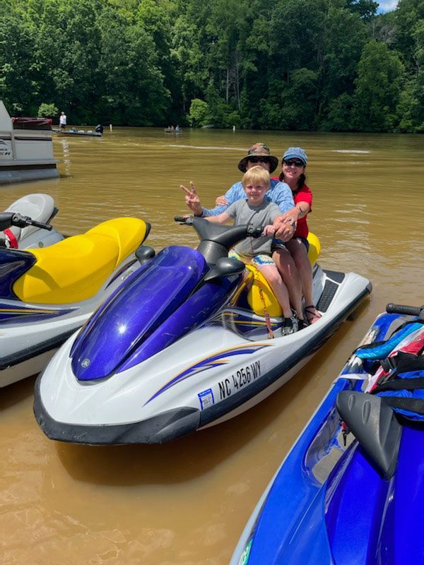 Three people riding a purple and white jet ski on a muddy river, with trees in the background.