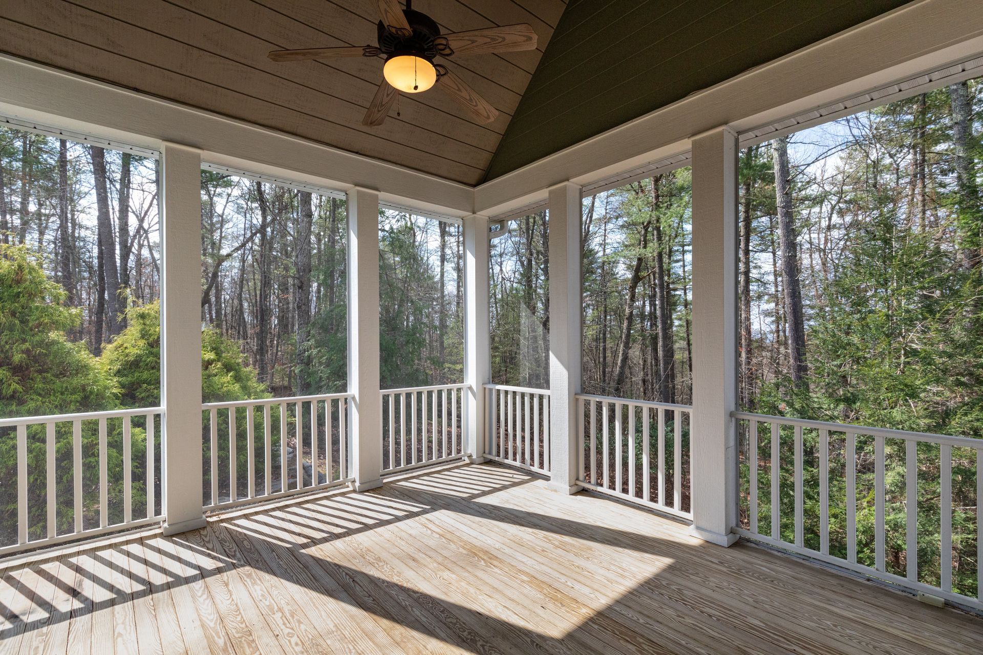 Covered porch overlooking wooded backyard with white railings and sunlit wooden floorboards.